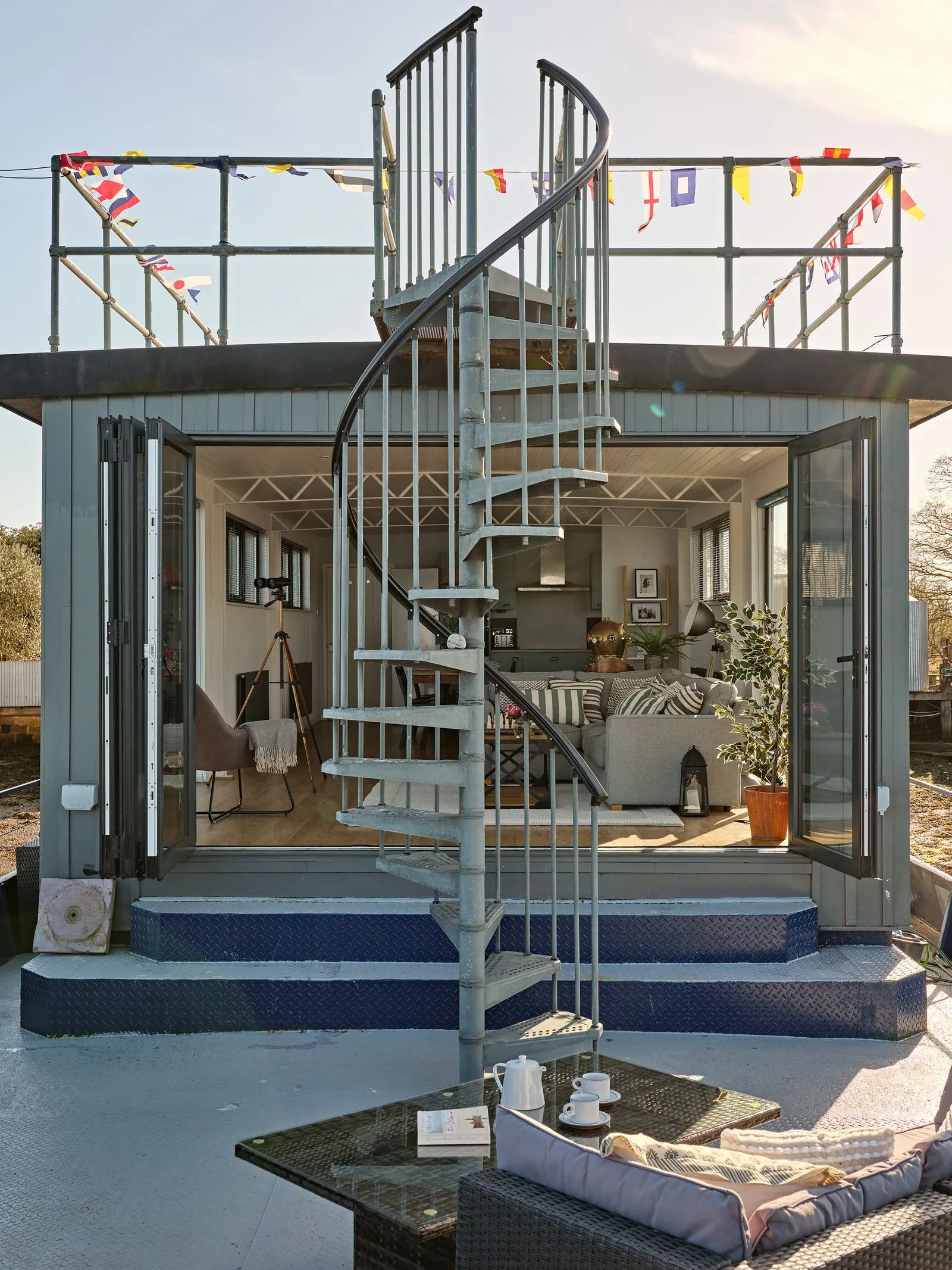 Modern tiny house with open glass doors, a spiral staircase, and outdoor seating with a coffee set on a glass table.