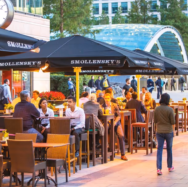 People dining at outdoor tables under Smollensky's black umbrellas in a city plaza during daylight.