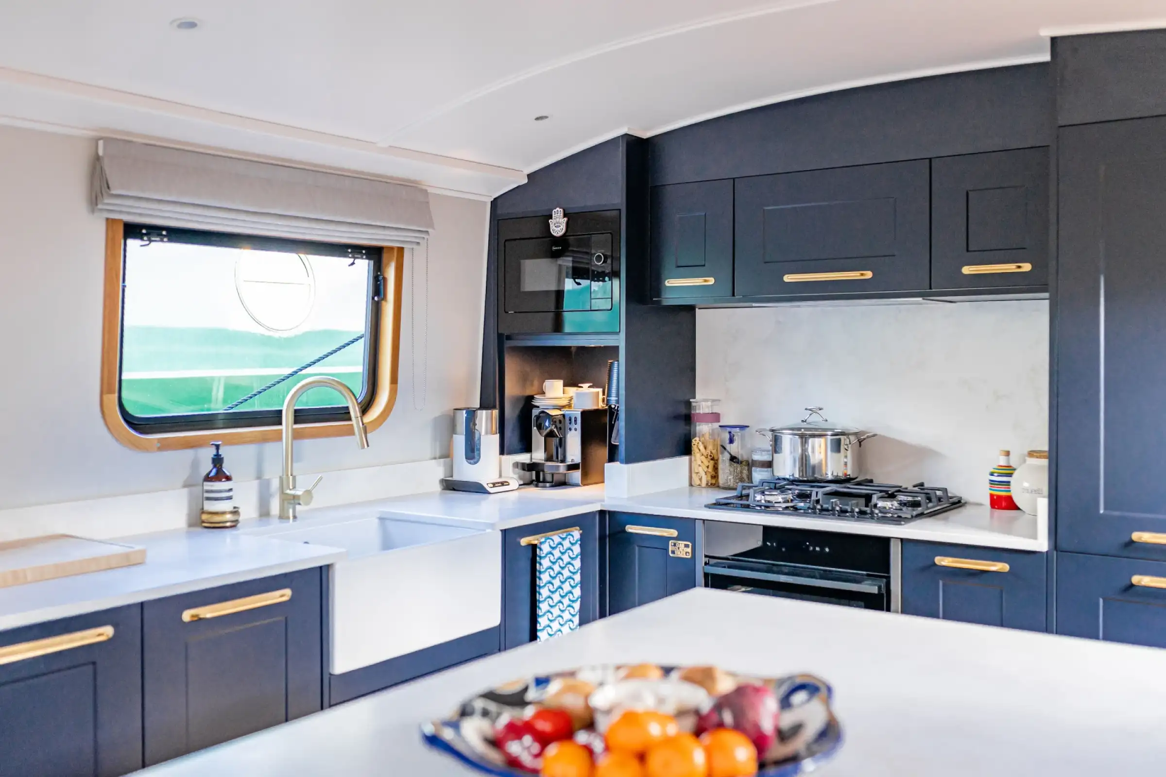 Modern kitchen with navy blue cabinets, gold handles, a white farmhouse sink, built-in microwave, gas stove with pot, and a window above the sink.
