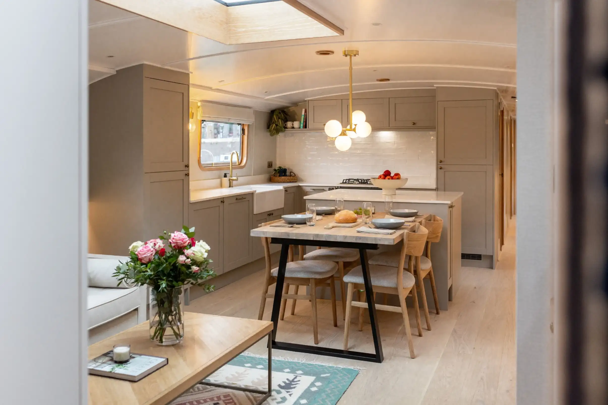 Modern kitchen and dining area with light wood furniture, gray cabinets, a vase with flowers, and a dining table set with dishes and a loaf of bread.