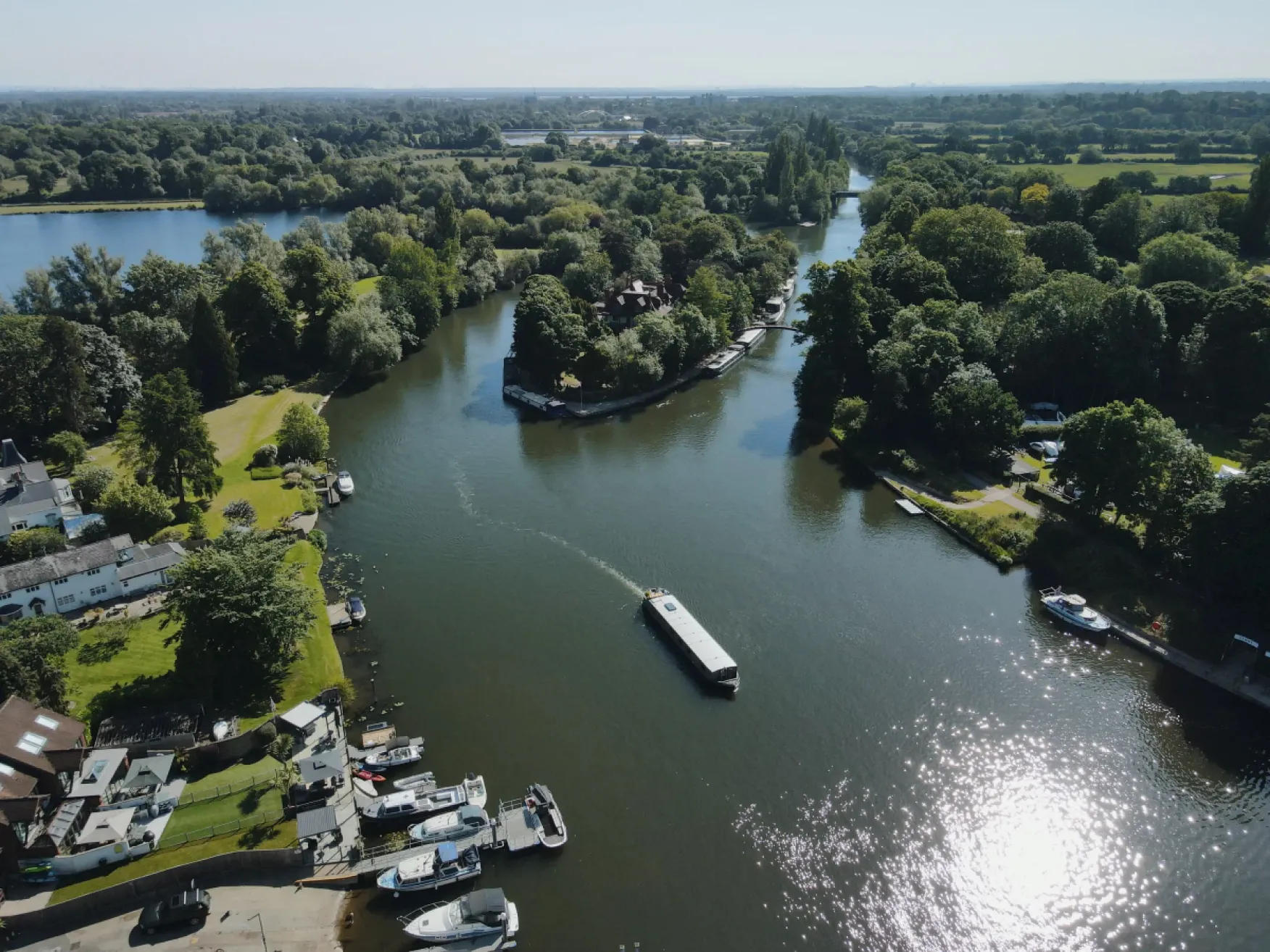 A river bends around a tree-covered island with boats docked along the shore on a sunny day.