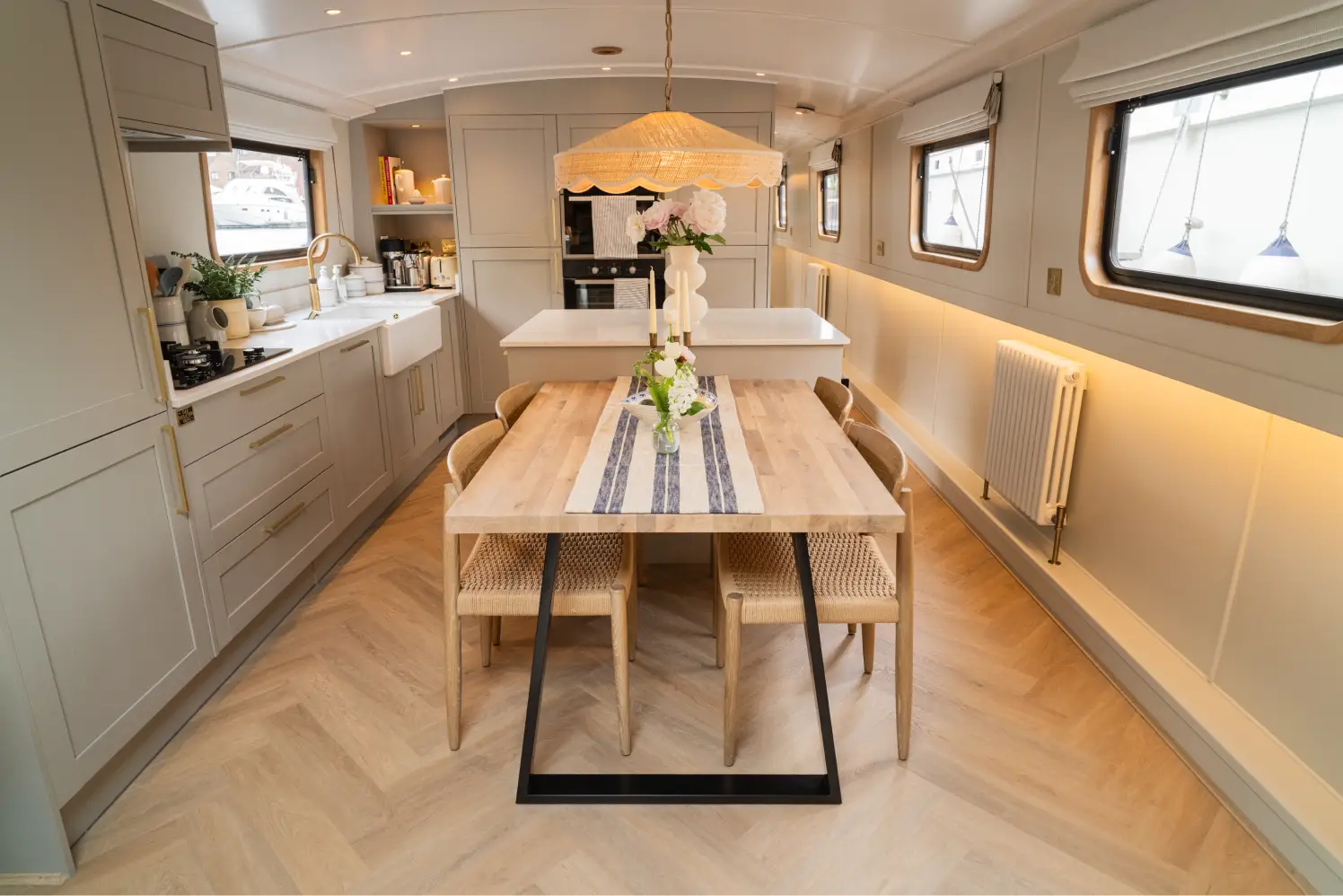 Cozy boat interior with a dining table set for four with wine glasses and a bottle, leather sofa, armchair, and windows showing water outside.