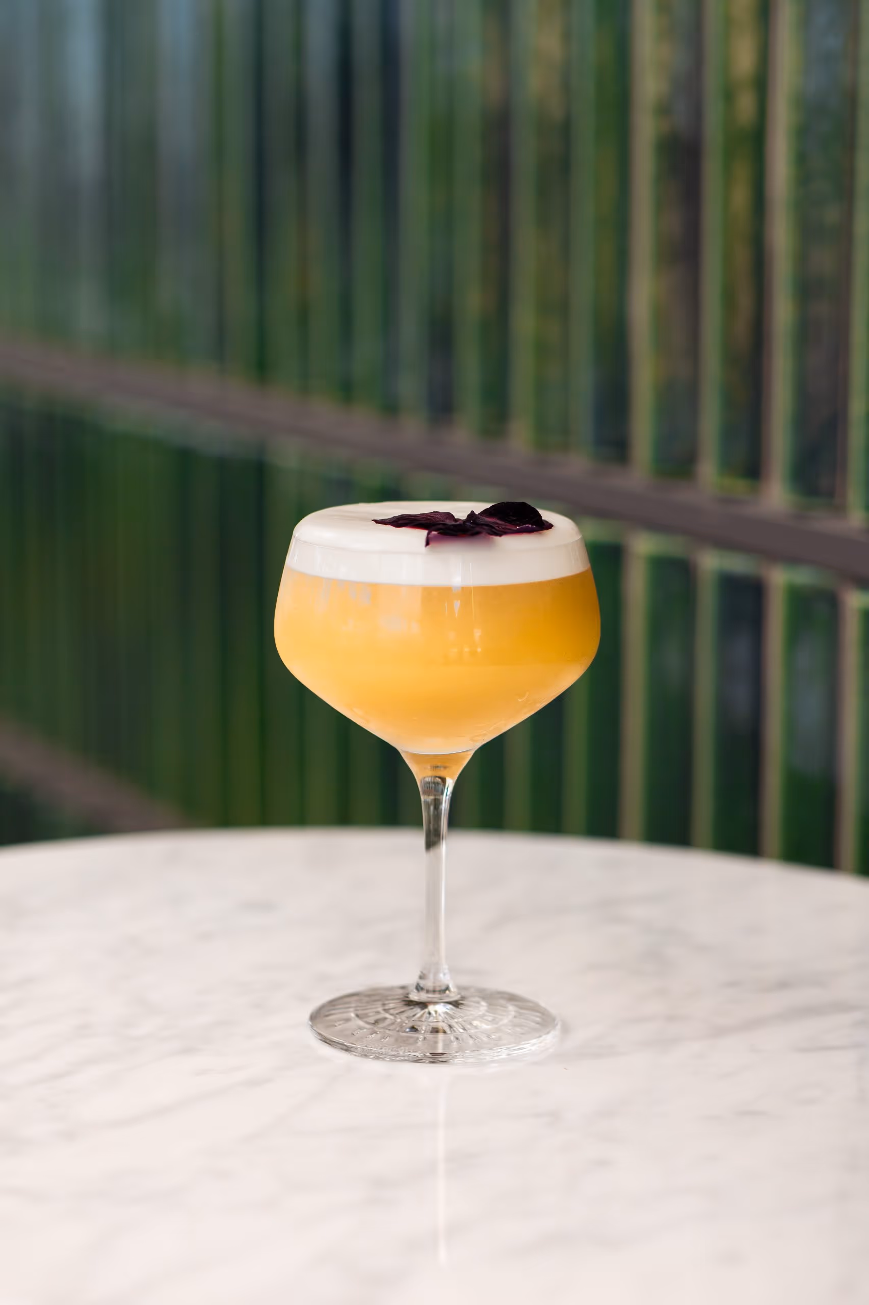 Yellow cocktail with white foam and purple garnish in a stemmed glass on a marble table.