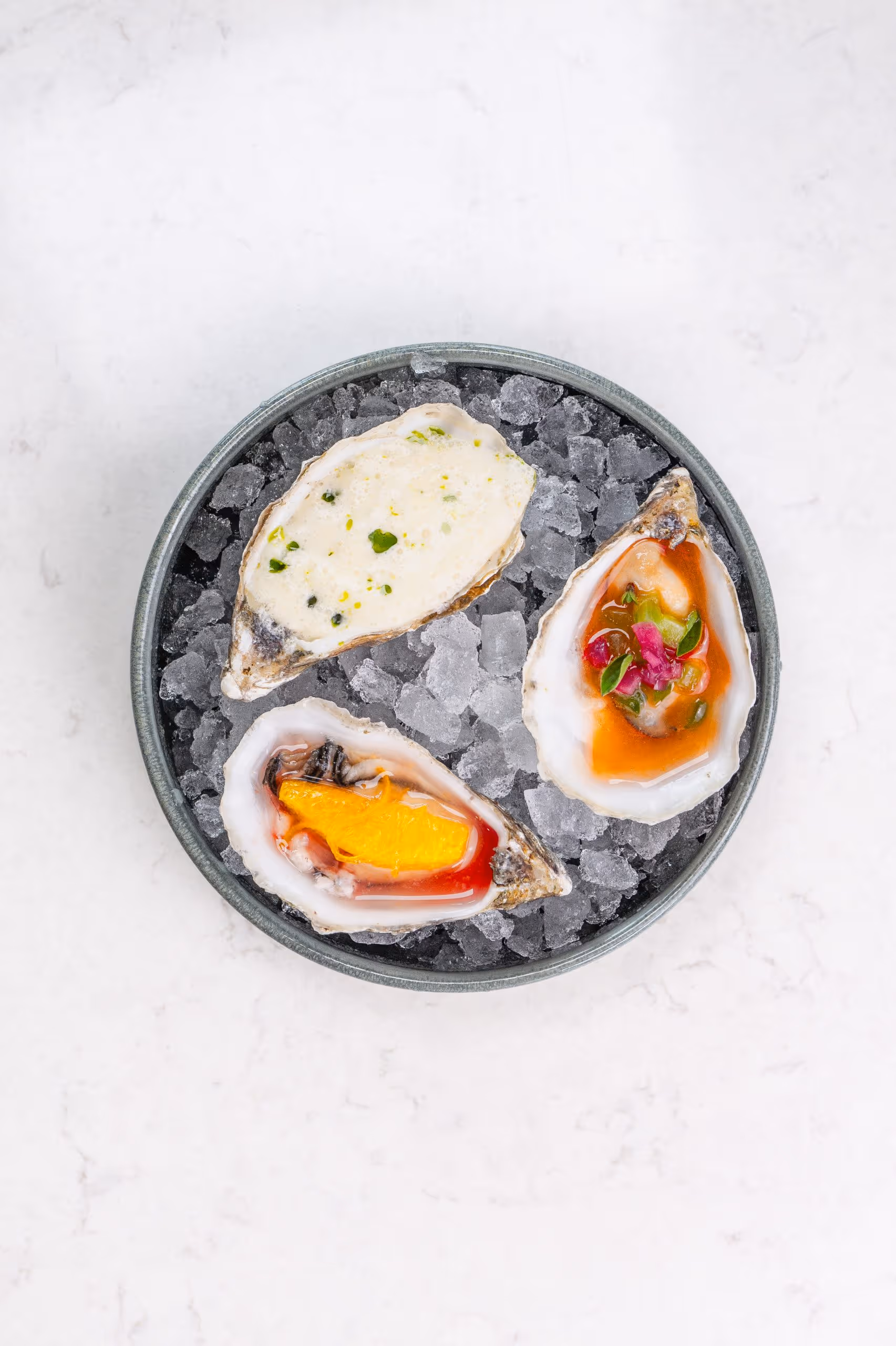 Three oysters served on crushed ice in a round dish, each topped with different garnishes including sauce, herbs, and a slice of orange.