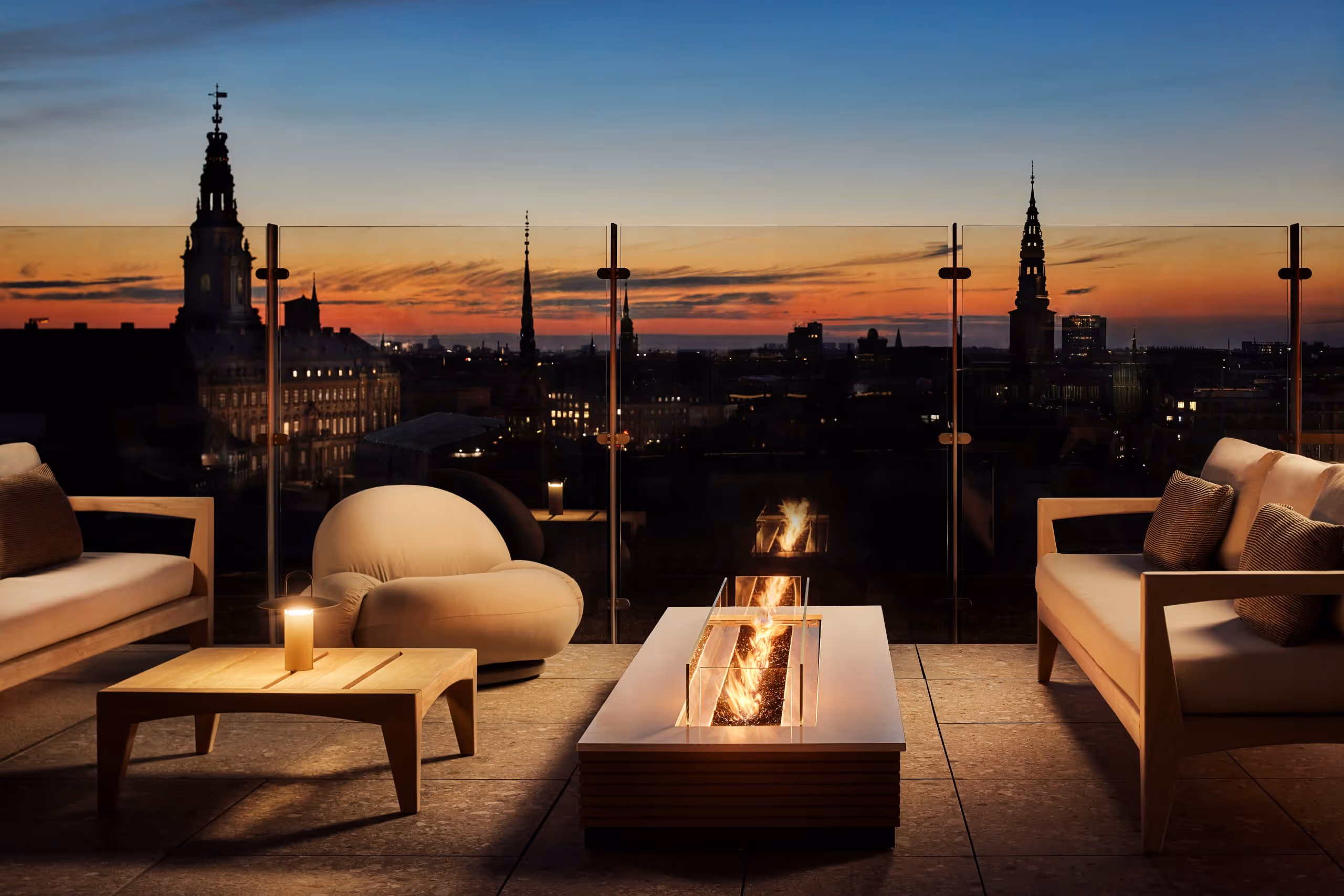 Modern rooftop terrace with cozy seating, fire pit, and city skyline at sunset.