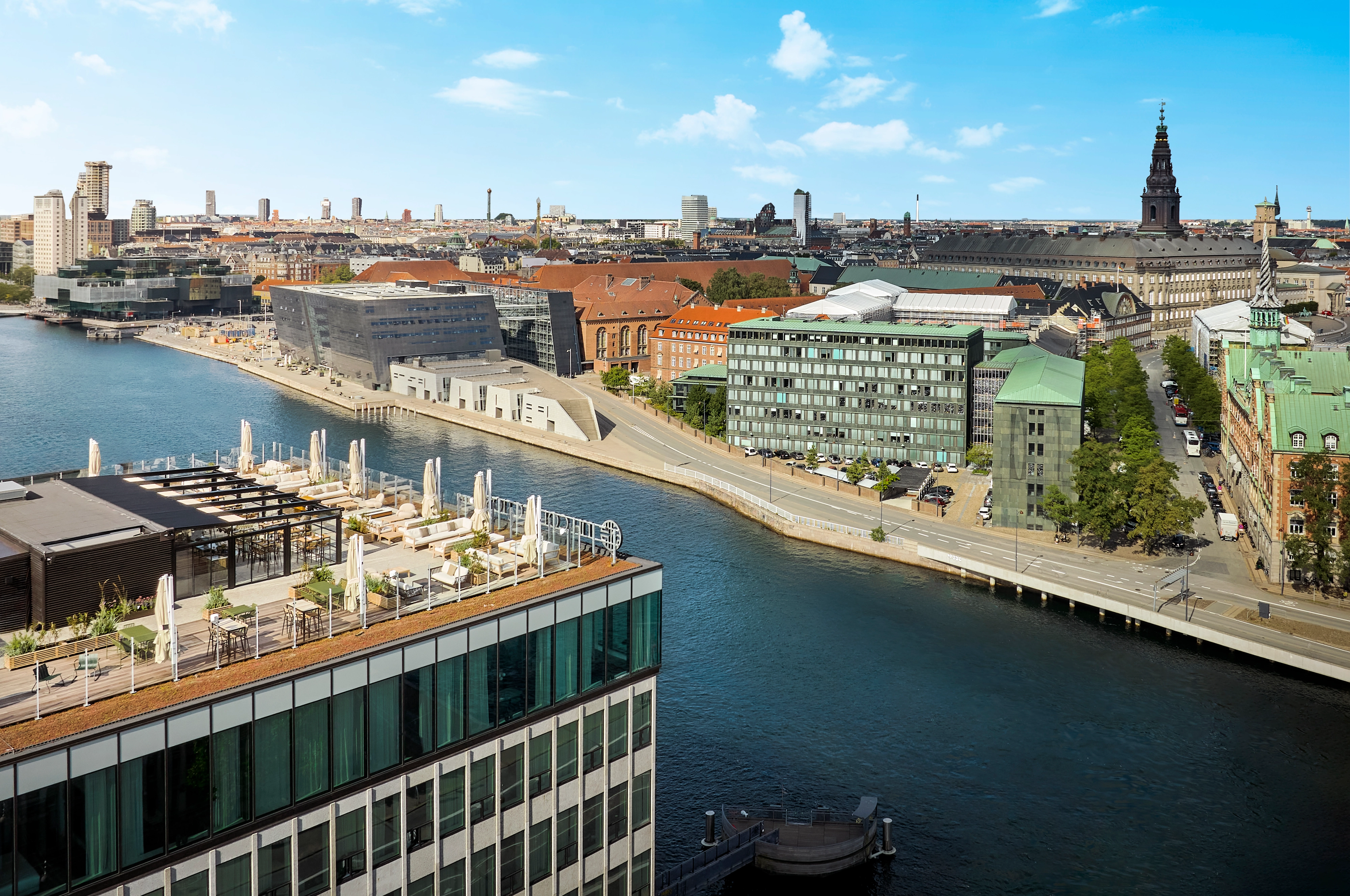 Aerial view of a modern rooftop terrace with seating and umbrellas overlooking a canal in Copenhagen, with historic and contemporary buildings in the background under a blue sky.