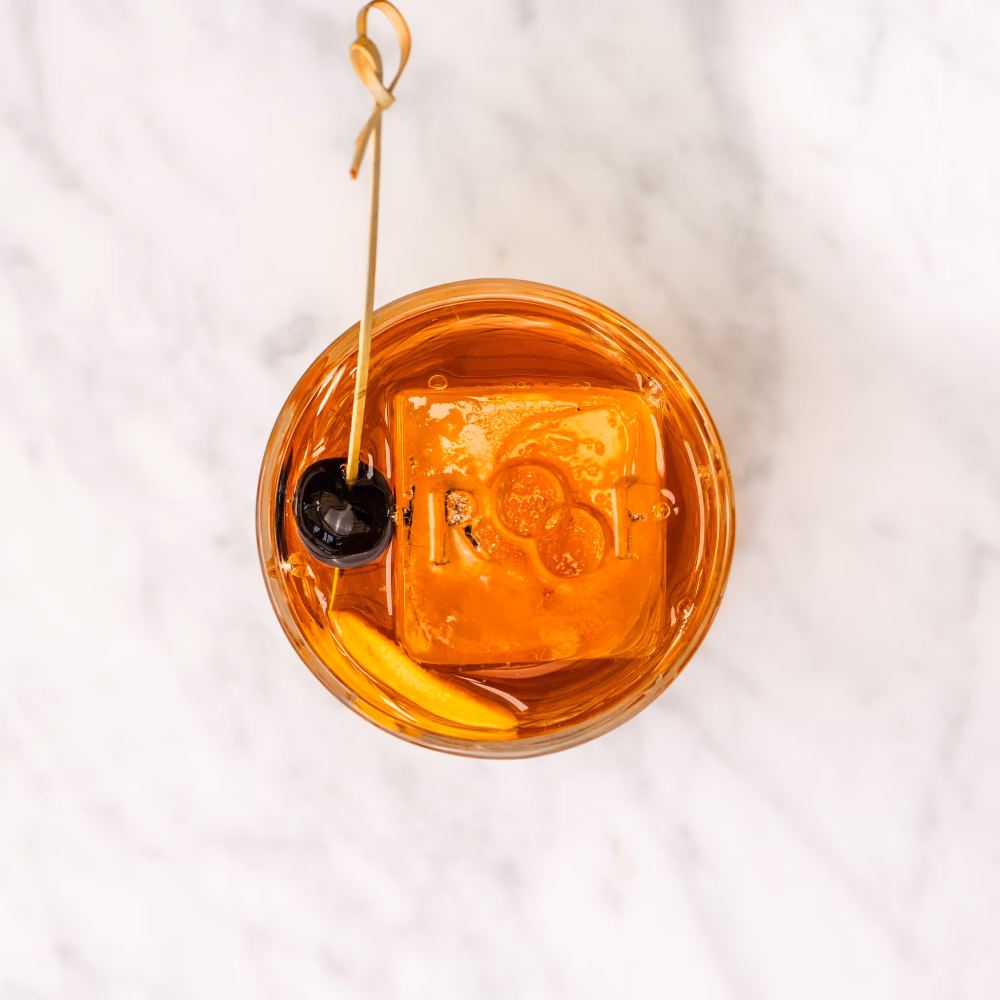 Top view of an amber cocktail with a large ice cube engraved with the letters ROF, garnished with a black cherry and an orange peel on a marble surface.
