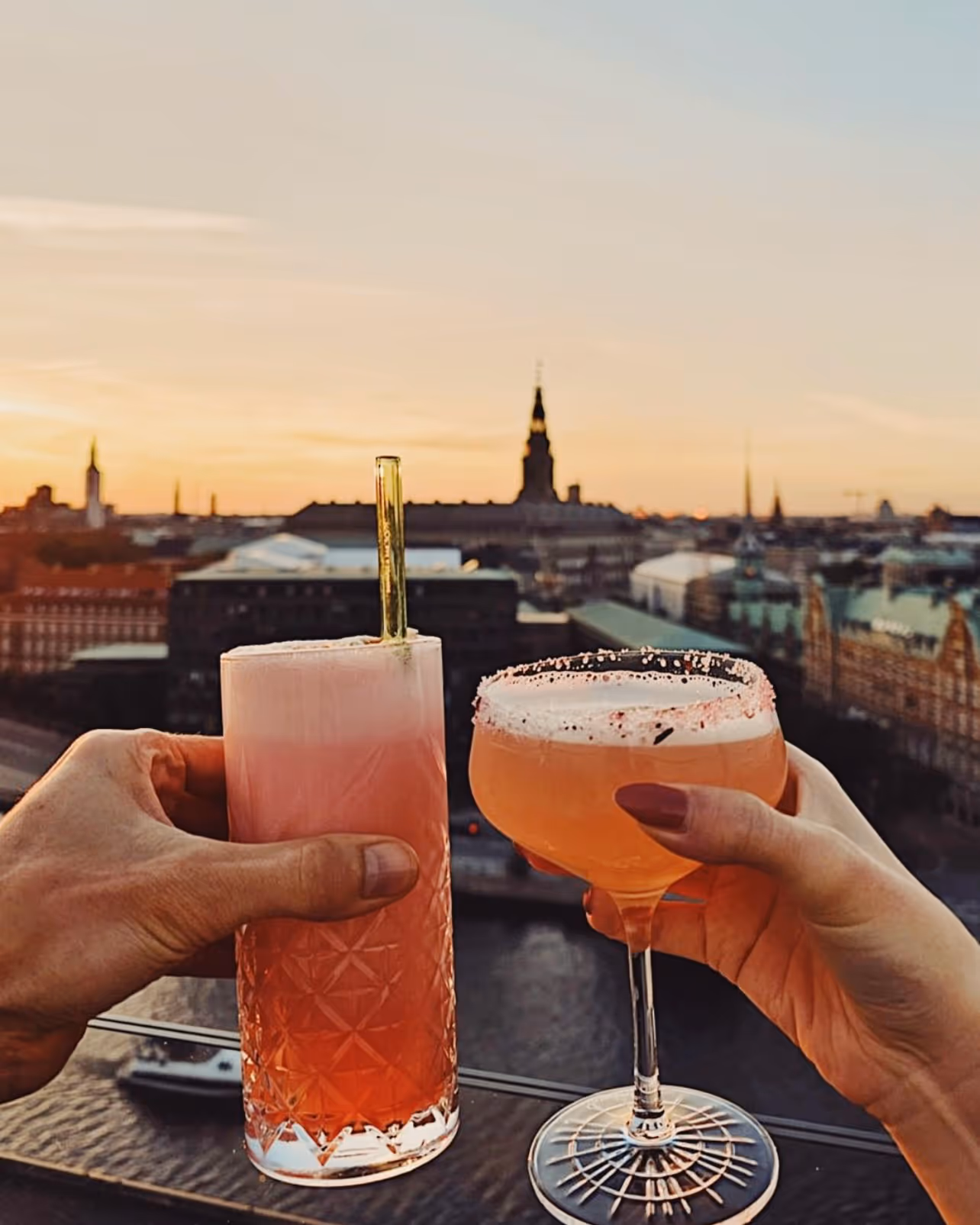 Two hands holding pink cocktails against a cityscape at sunset, with water and historic buildings in the background.
