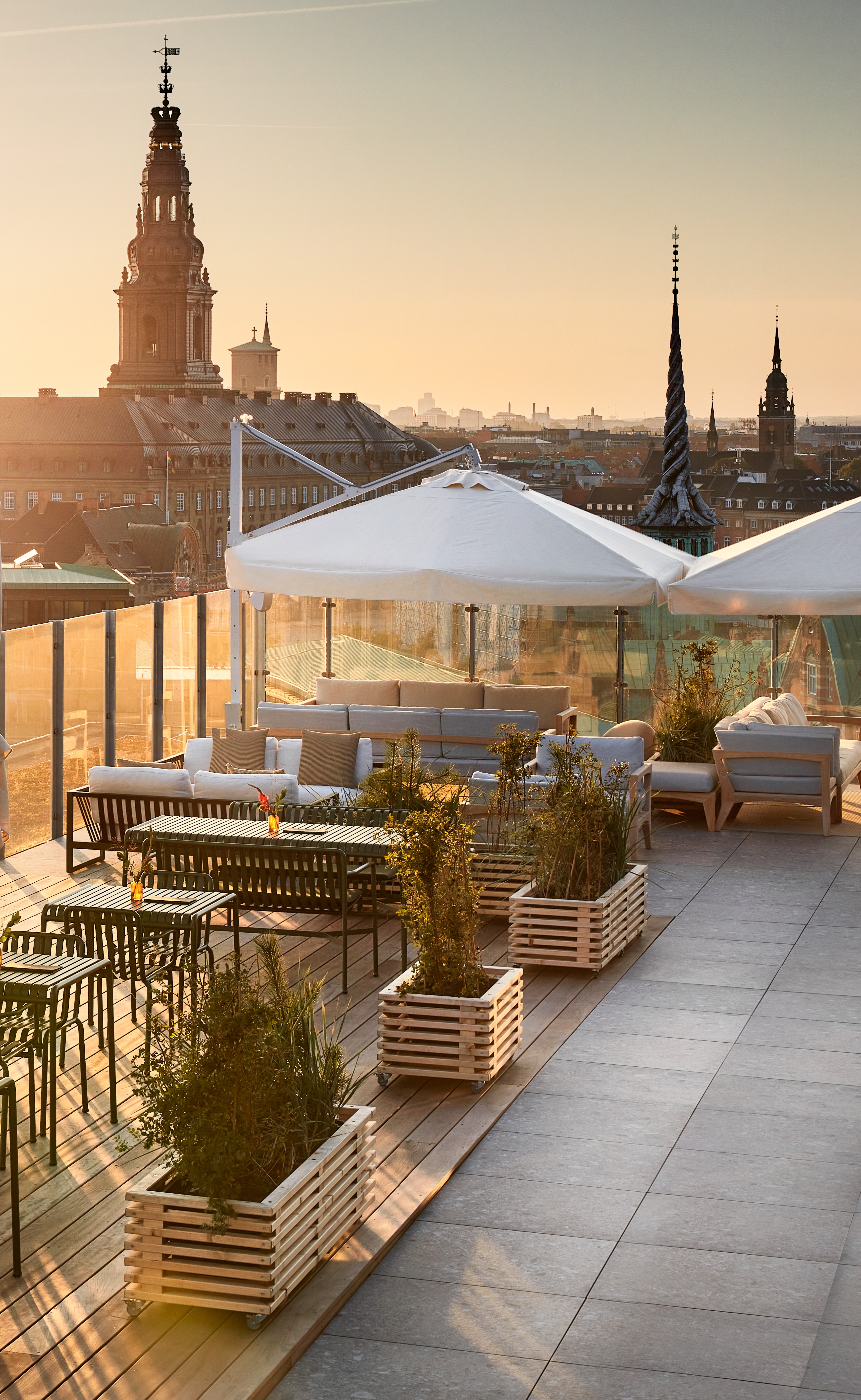 Rooftop terrace with outdoor seating, white umbrellas, and potted plants overlooking a city skyline at sunset.