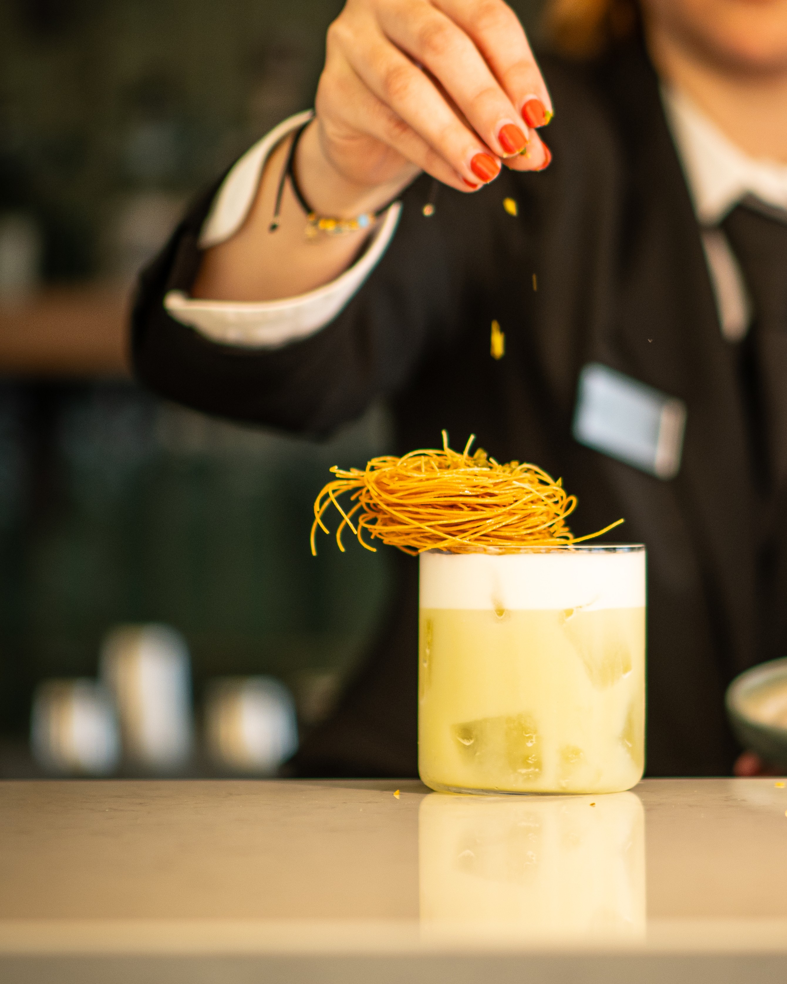 Hand with red nail polish sprinkling garnish onto a frothy iced cocktail topped with a nest of crispy noodles.
