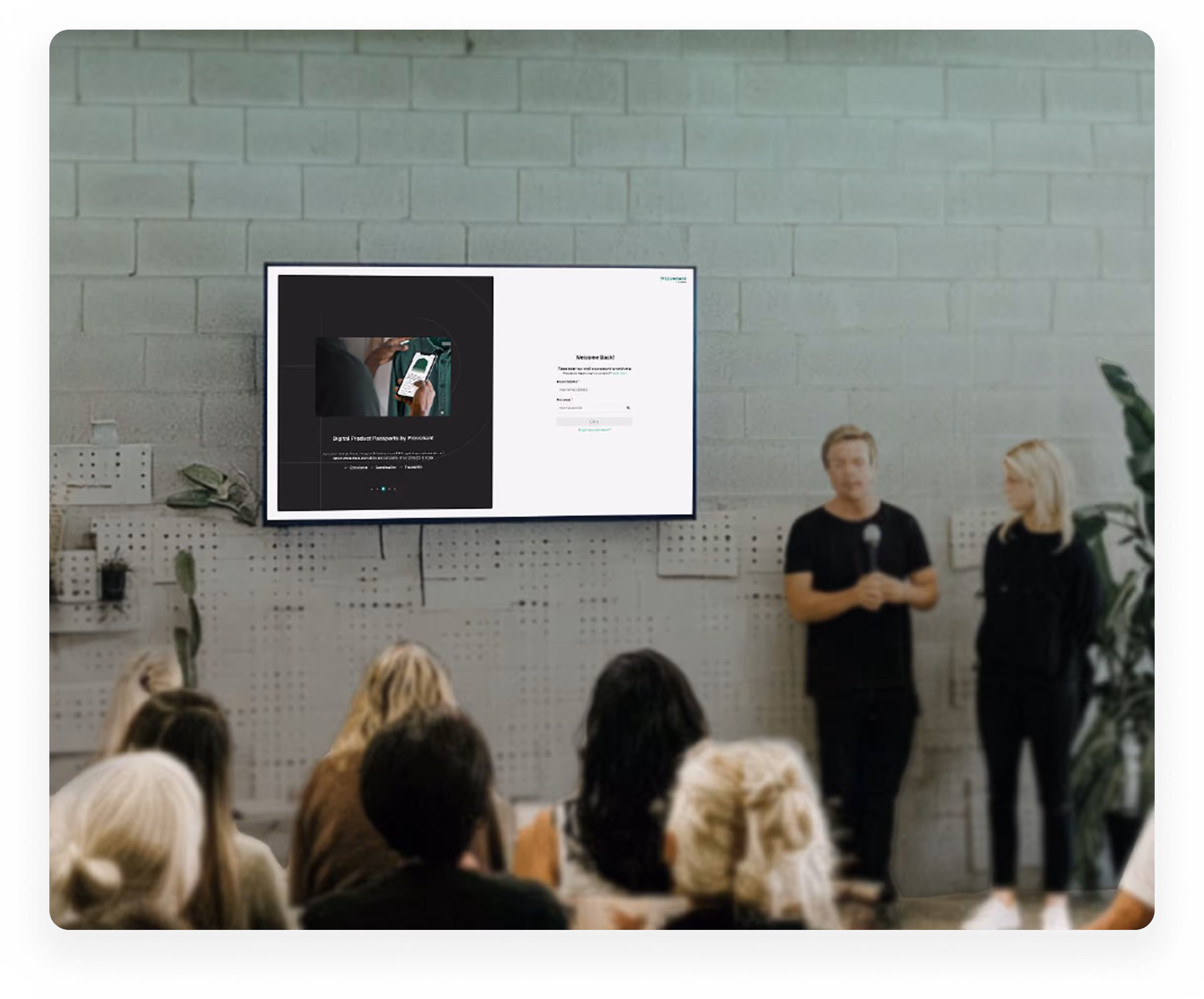 Two presenters standing next to a screen displaying a digital product login page, facing an audience in a room with gray brick walls.
