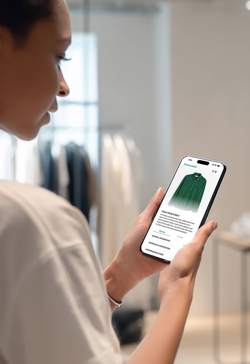 Person holding a smartphone displaying a product page for a green oxford shirt in a clothing store.