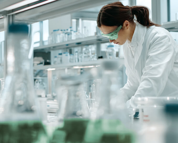 Scientist wearing green safety goggles and a white lab coat working in a laboratory with glassware and chemical containers.