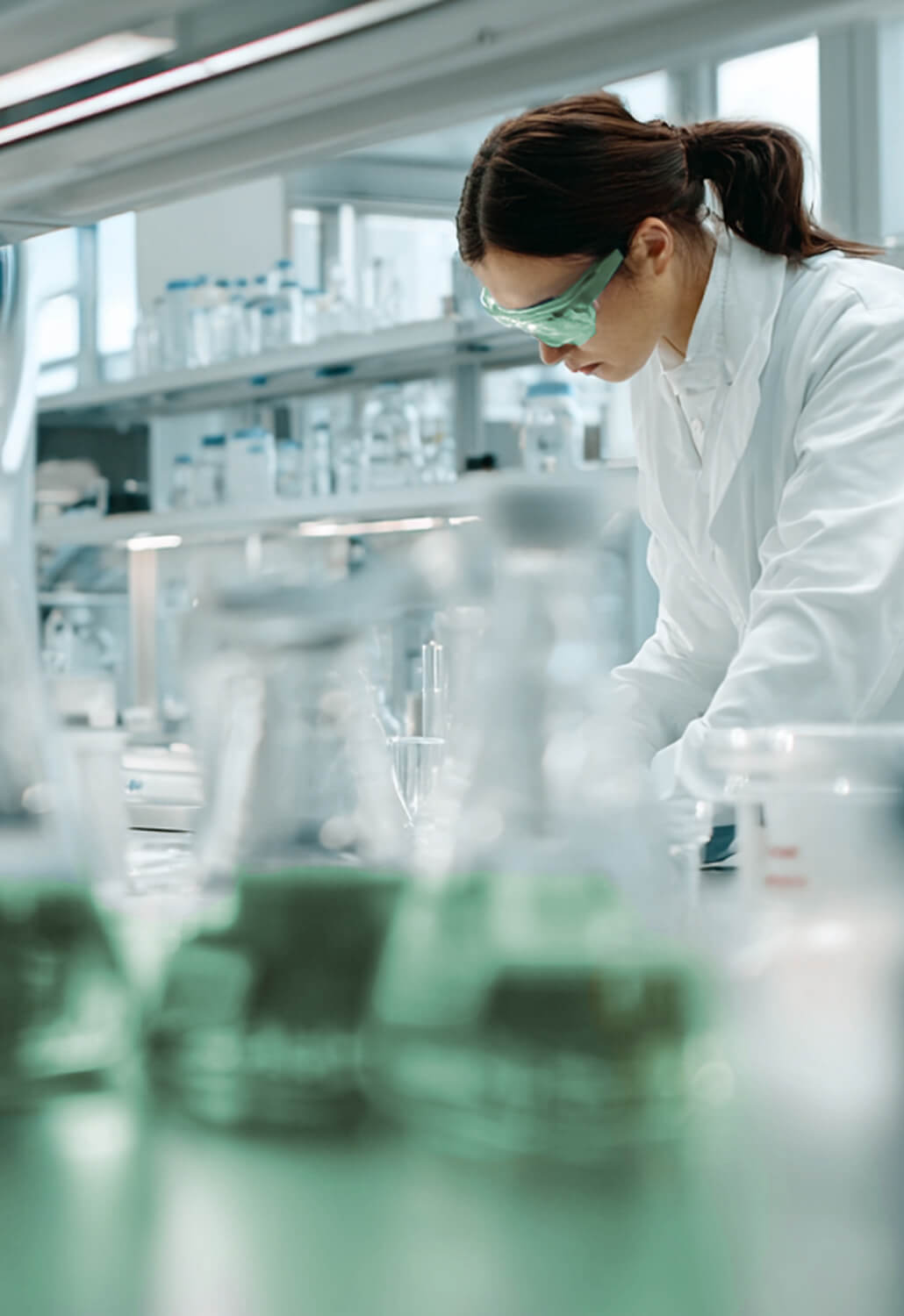 Female scientist wearing green safety goggles and a white lab coat working in a laboratory with glass beakers and equipment.