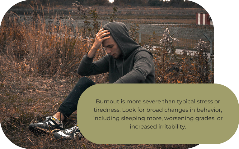 Image: A teenager in a gray hoodie sitting in a brown field of tall grass with his hand n his forehead. Text: Burnout is more severe than typical stress or tiredness. Look for broad changes in behavior, including sleeping more, worsening grades, or increased irritability. 