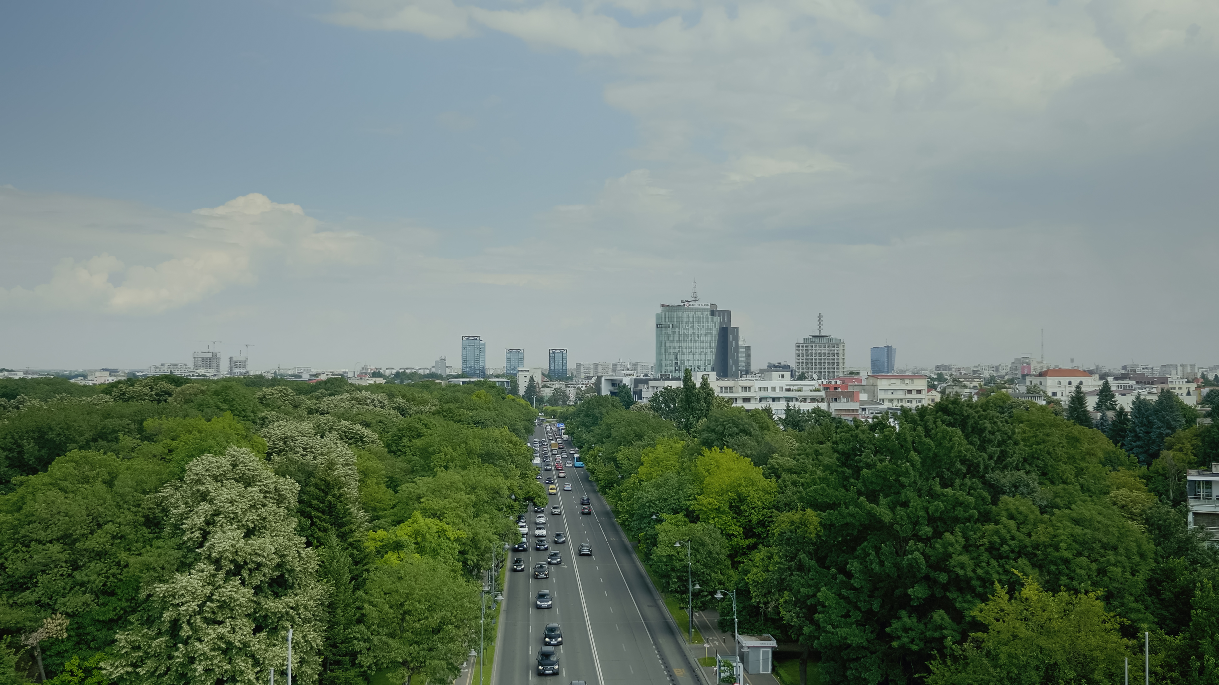Aerial view of a city with a busy road flanked by dense green trees and modern buildings in the background under a partly cloudy sky.