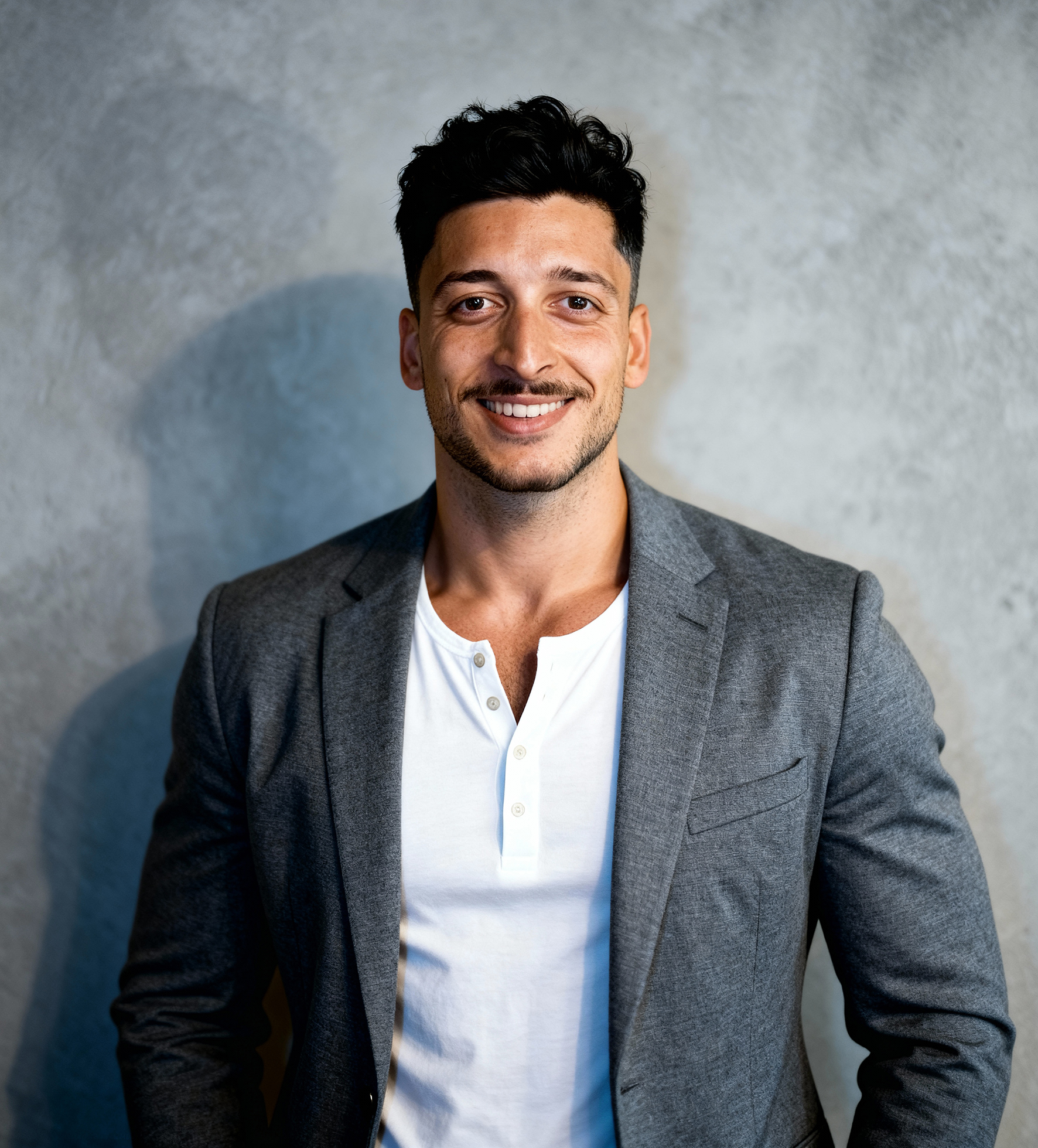 Smiling man with dark hair wearing a gray blazer and white henley shirt standing against a textured gray wall.