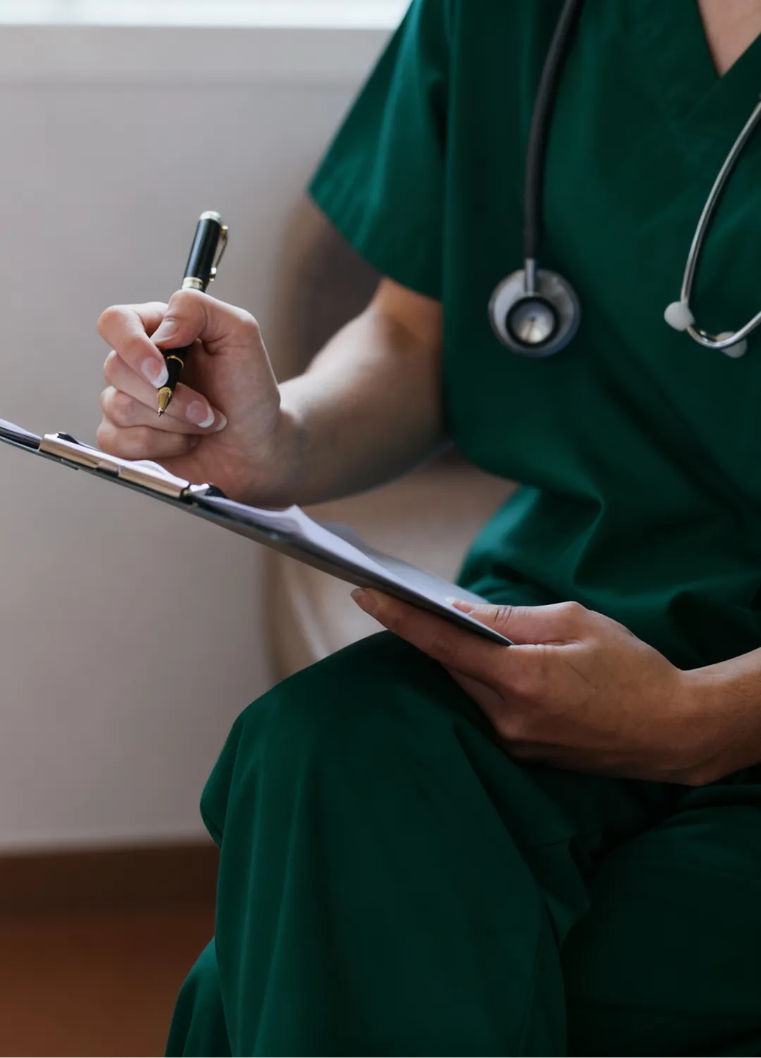 Medical professional in green scrubs with a stethoscope writing notes on a clipboard.