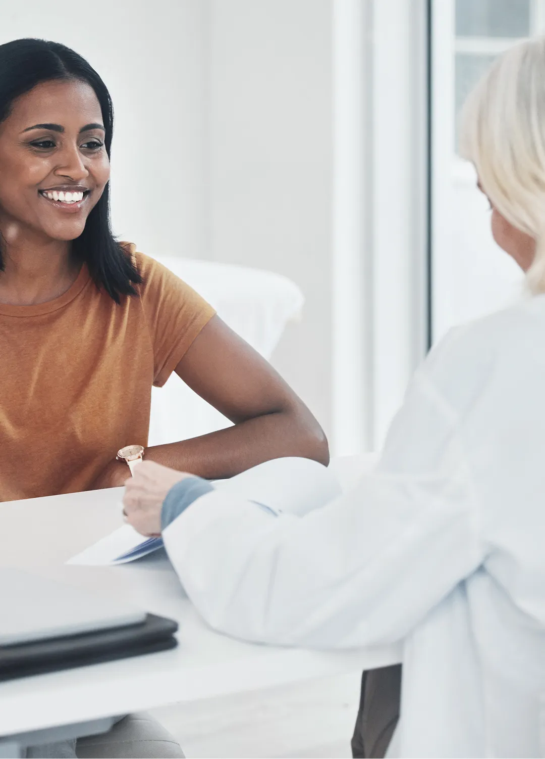 Smiling woman in orange shirt talking to a healthcare professional holding a clipboard in a bright office.