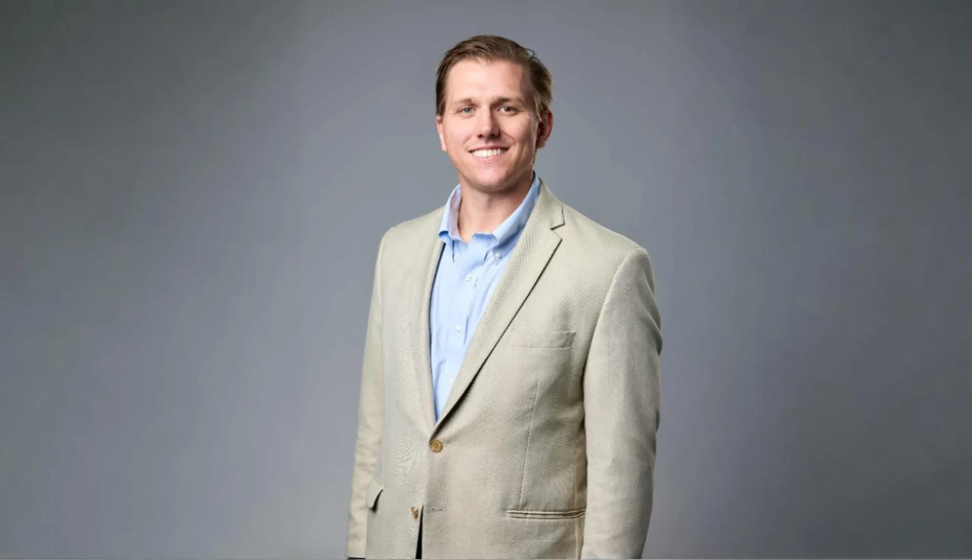 Smiling man wearing a beige blazer and light blue shirt standing against a gray background.