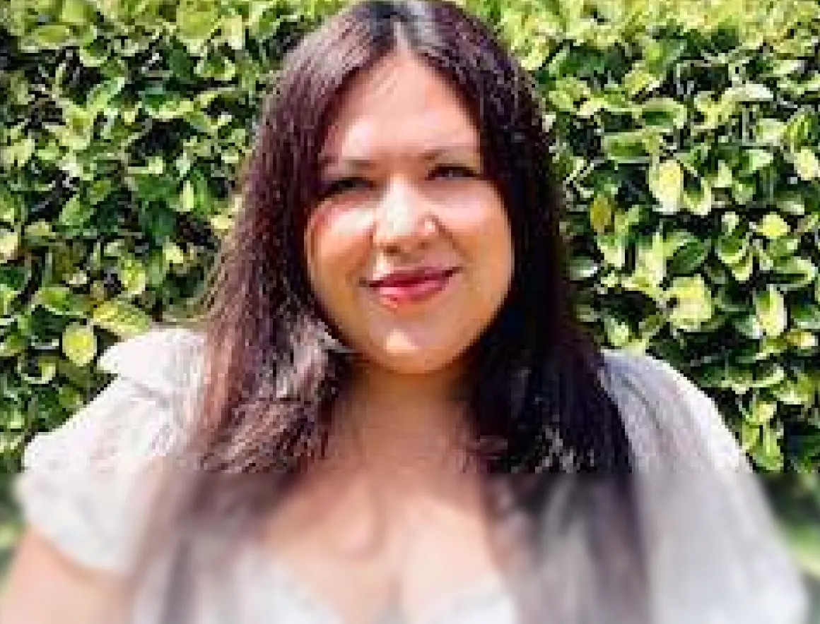 Smiling woman with long dark hair standing outdoors in front of green leafy bushes.