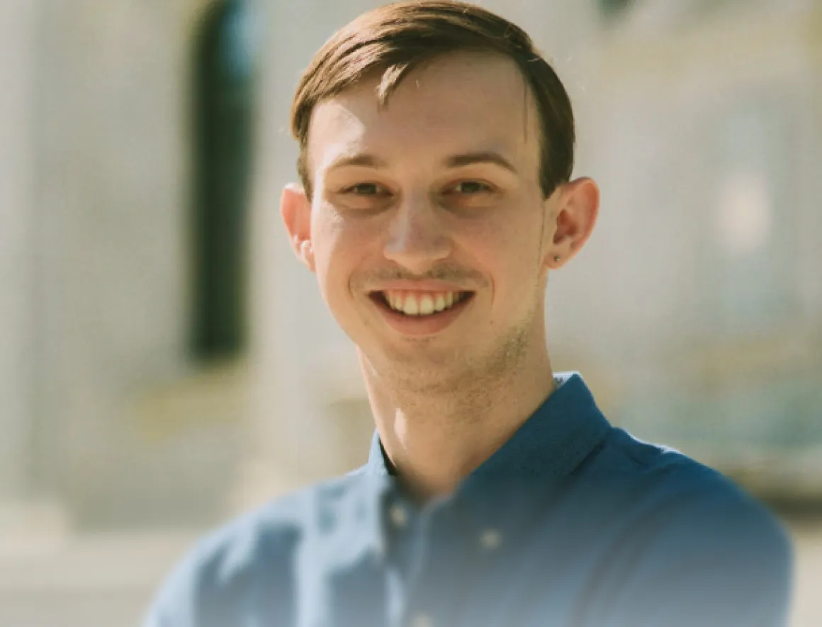 Smiling young man with short brown hair wearing a blue button-up shirt in an outdoor setting.