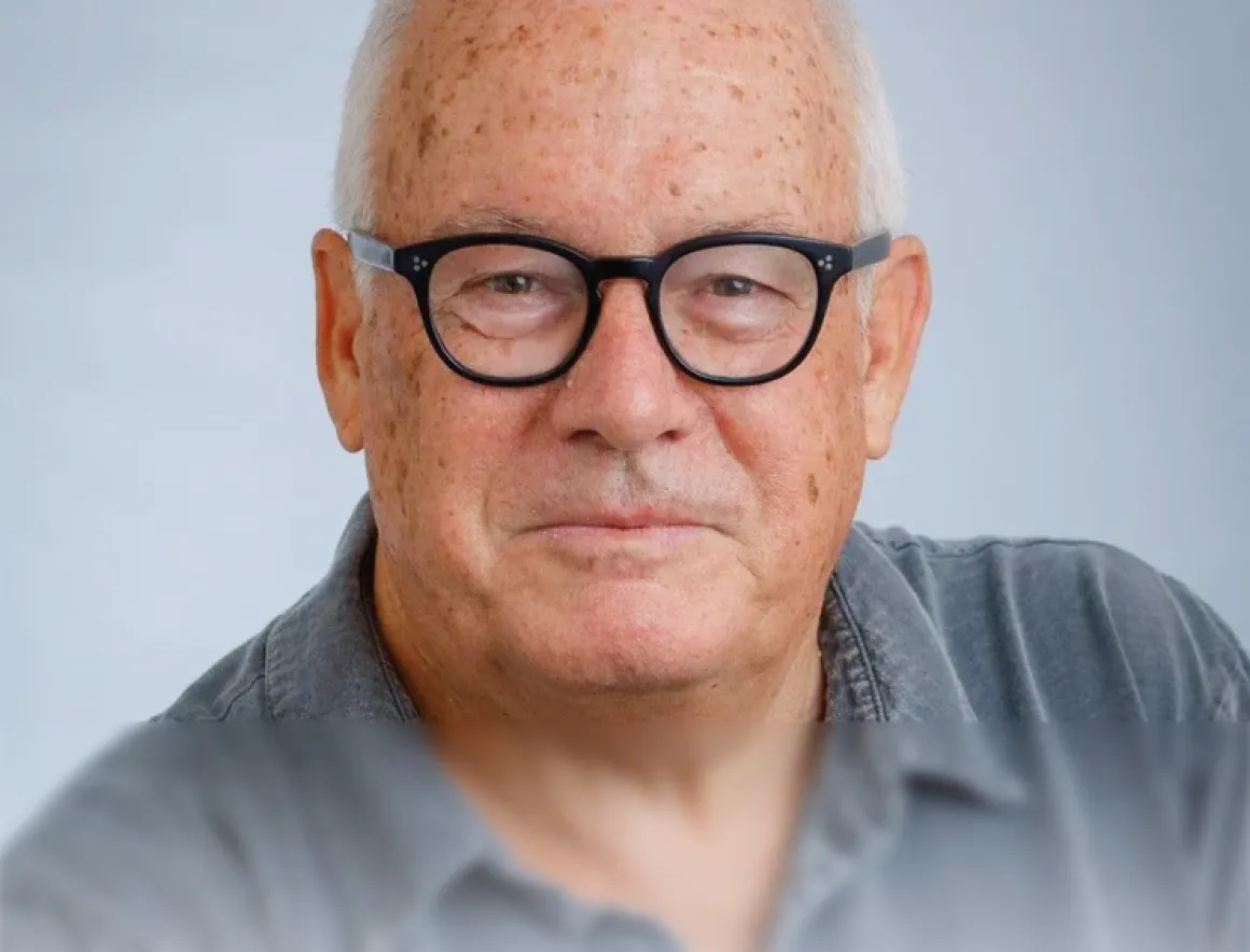 Close-up portrait of an elderly man with white hair and black glasses wearing a gray shirt.