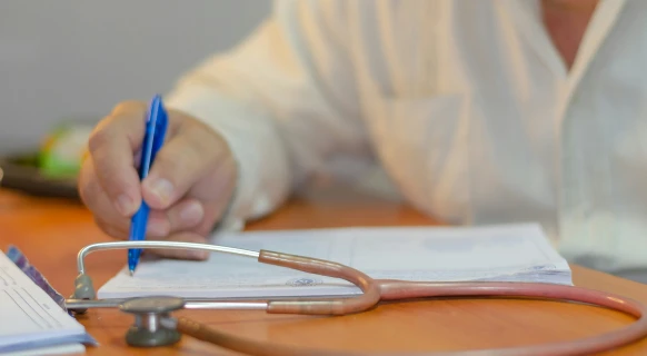 Person in a light shirt writing with a blue pen near a stethoscope on a wooden table.
