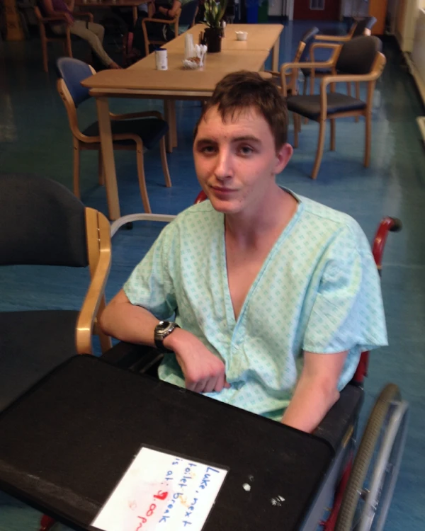 Young man in a wheelchair wearing a hospital gown sitting at a table in a room with chairs and a larger table in the background.
