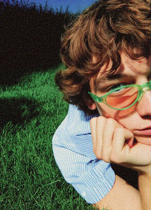 Person lying on grass with head resting on hand wearing green-tinted sunglasses