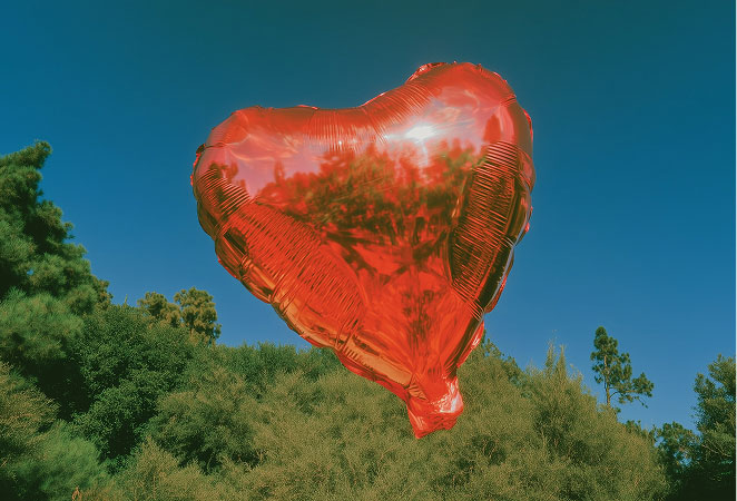 Red heart-shaped balloon floating in the sky above green trees