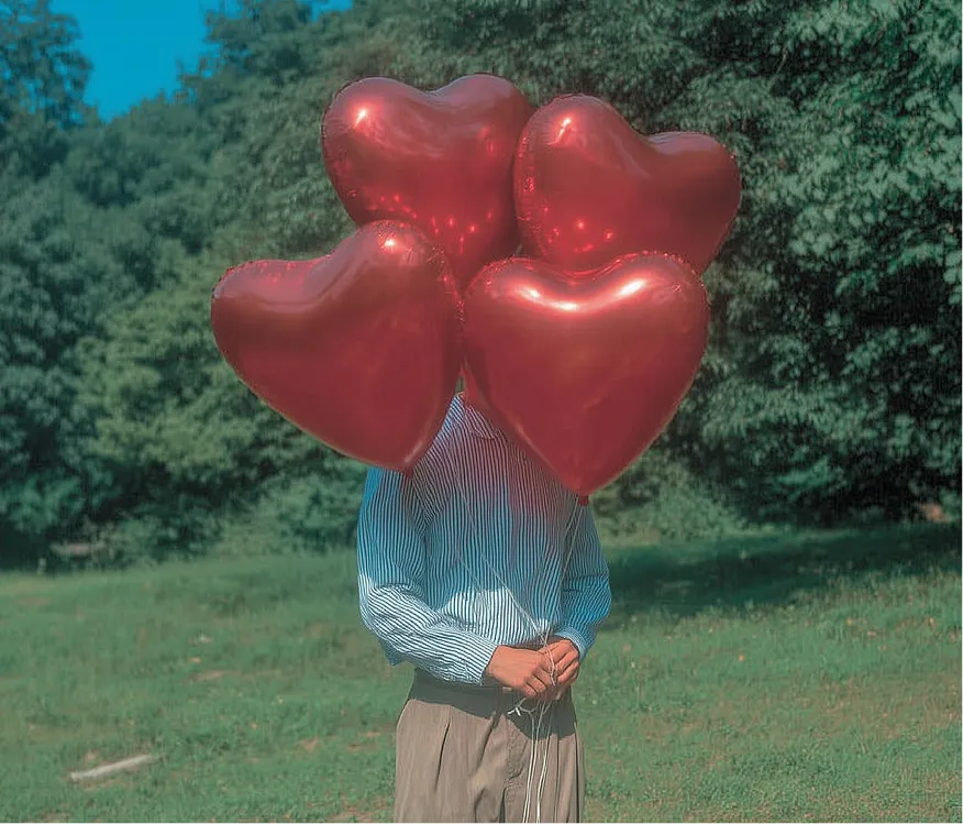 Person holding red heart-shaped balloons covering their face, standing in a green park on a sunny day