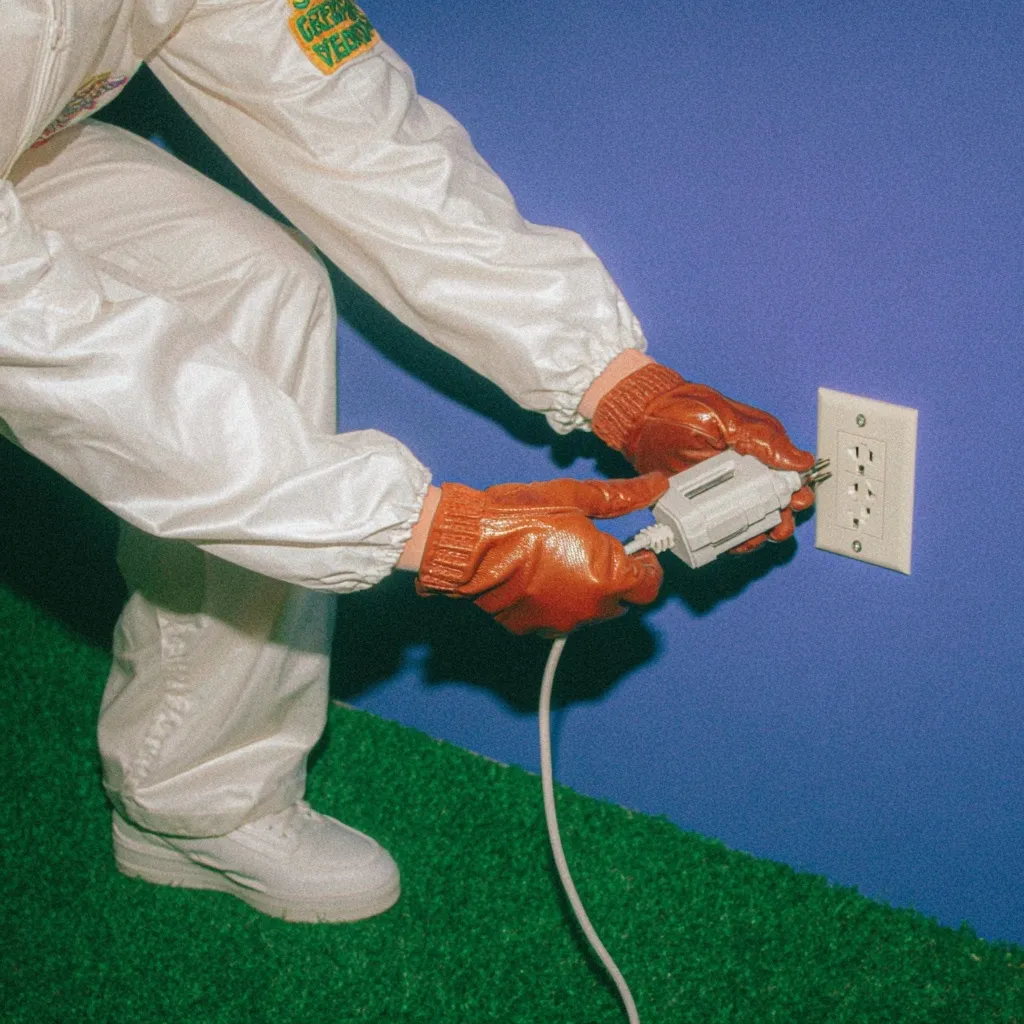 Person wearing white protective coveralls and brown gloves plugging a white electrical device into a wall outlet on a blue wall, standing on green carpet.
