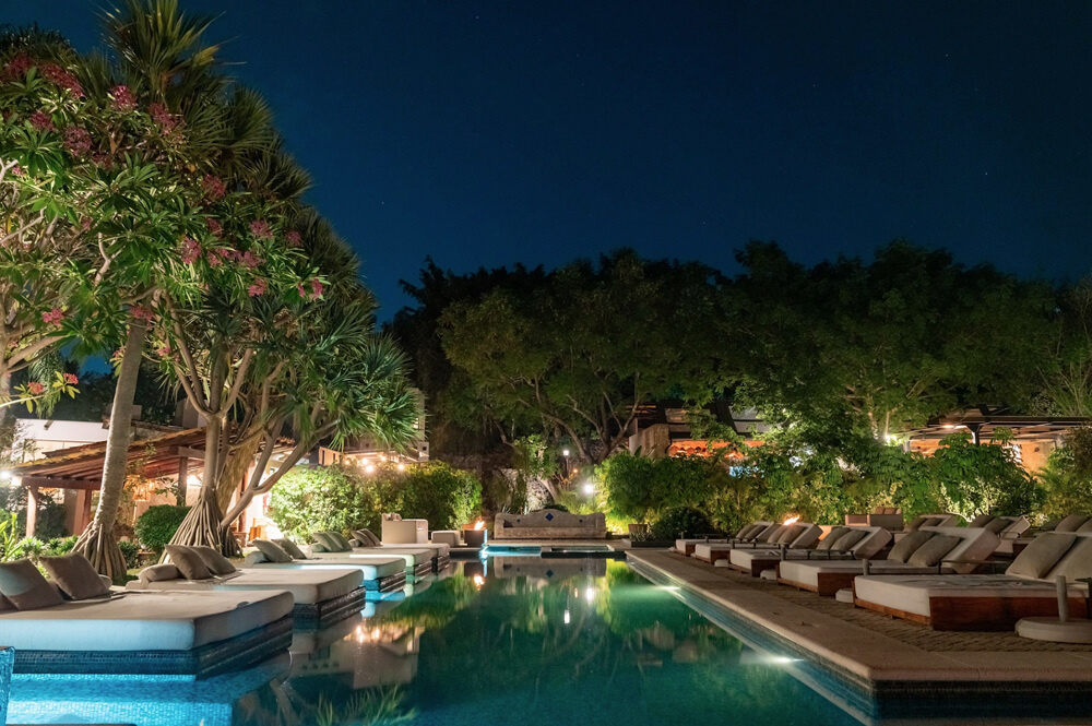 Piscina iluminada de noche rodeada de tumbonas y árboles verdes en un ambiente tropical.