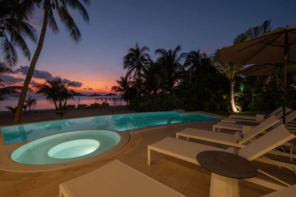 Piscina iluminada y jacuzzi en terraza con tumbonas y sombrillas, palmeras al atardecer junto a la playa.