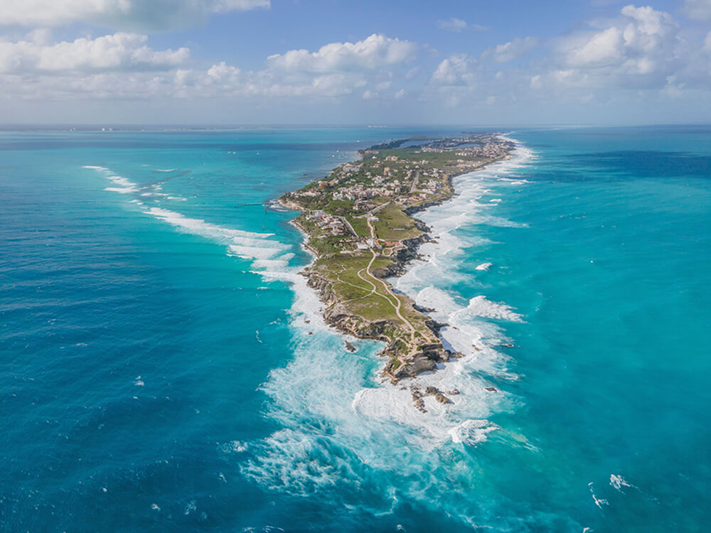 Vista aérea de una península estrecha con olas rompiendo en la costa y viviendas dispersas entre vegetación verde.