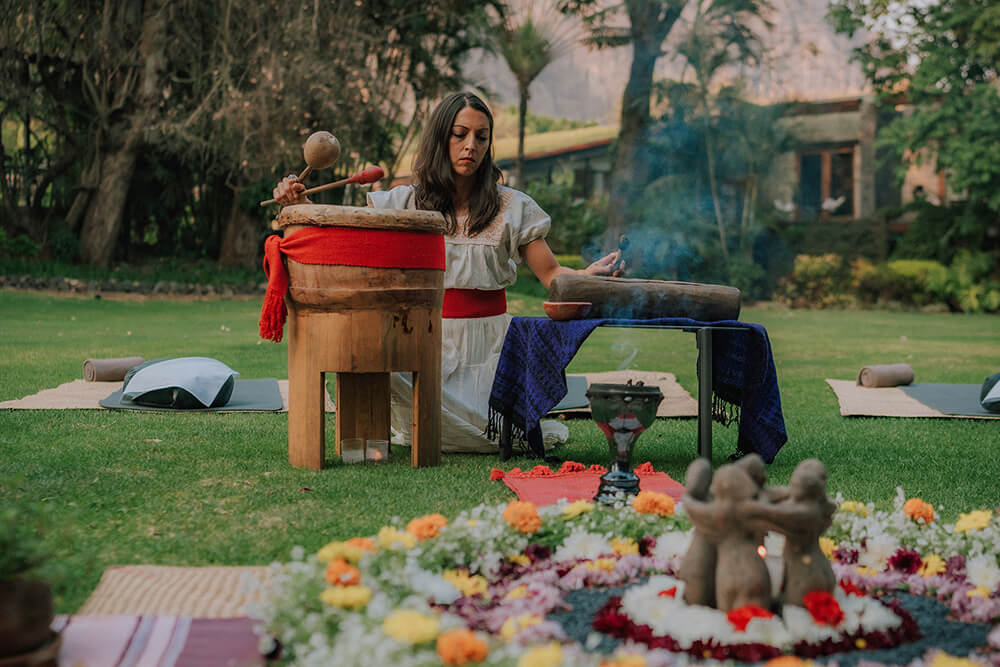 Mujer arrodillada en un jardín tocando un tambor grande con mazo, rodeada de arreglos florales y objetos ceremoniales.