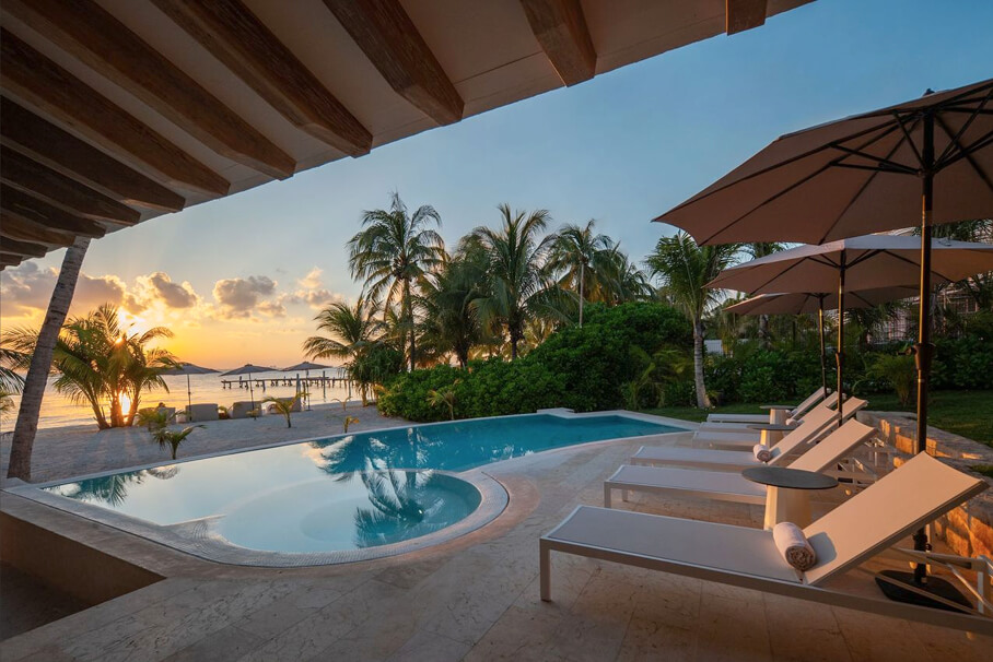 Piscina al aire libre con tumbonas y sombrillas, palmeras y playa al atardecer.