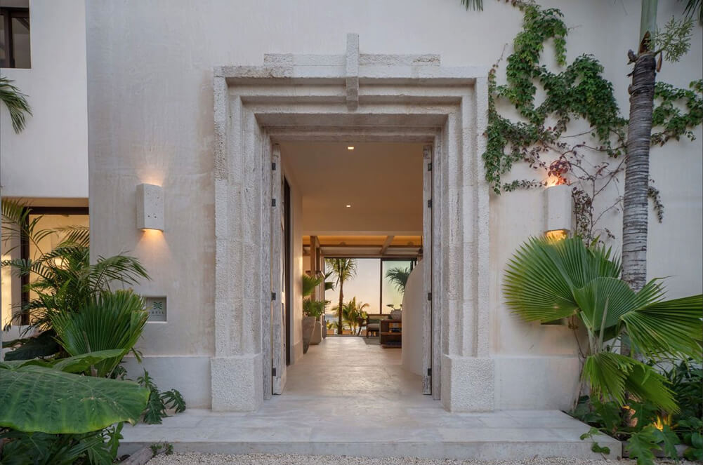 Entrada de casa con marco de piedra, paredes blancas y plantas tropicales alrededor.