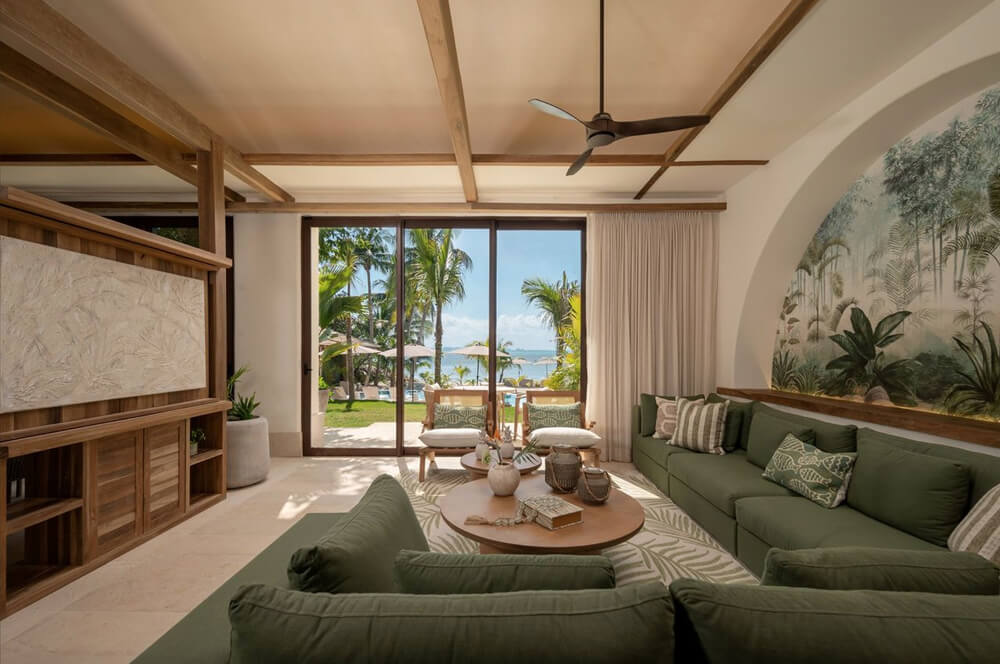 Sala de estar con sofás verdes, mesa de centro de madera y ventana grande que muestra palmeras y vista al mar.