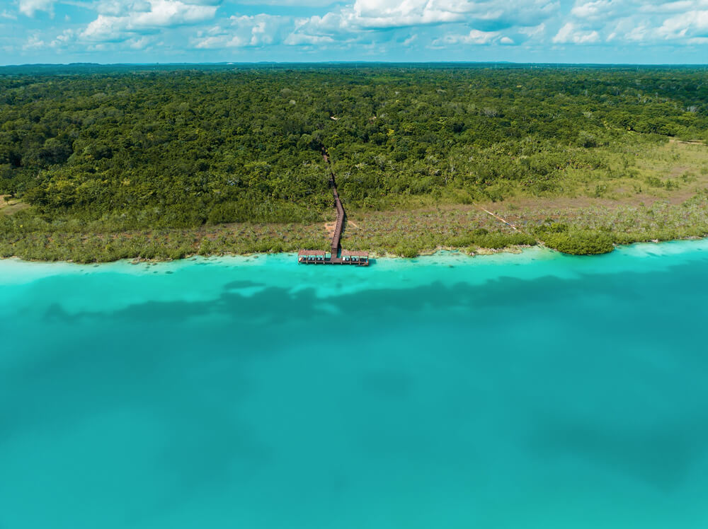Vista aérea de un muelle que se adentra en aguas turquesas frente a una densa costa de vegetación tropical.
