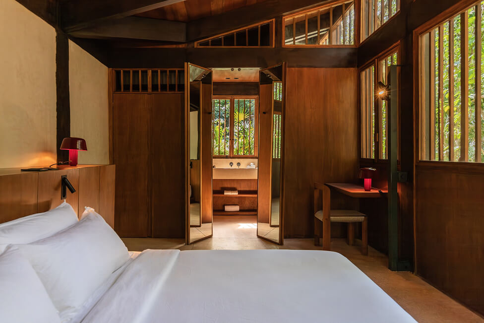 Habitación con cama blanca, paredes y muebles de madera, escritorio con lámpara roja y puerta abierta hacia baño con ventana que muestra vegetación.