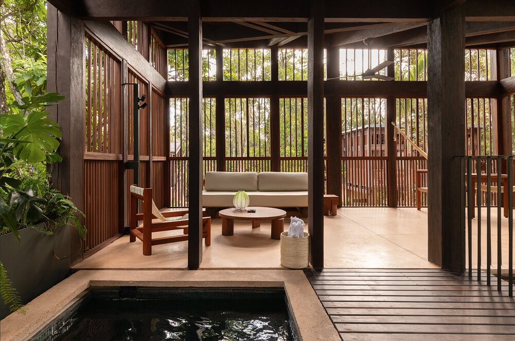 Sala al aire libre con paredes de listones de madera, sofá beige, mesa redonda baja y una silla de madera cerca de una piscina pequeña.