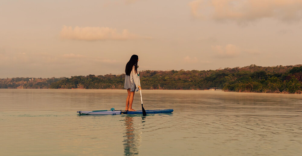 Persona de pie en una tabla de paddle surf en un lago tranquilo con un fondo de bosque y cielo nublado.