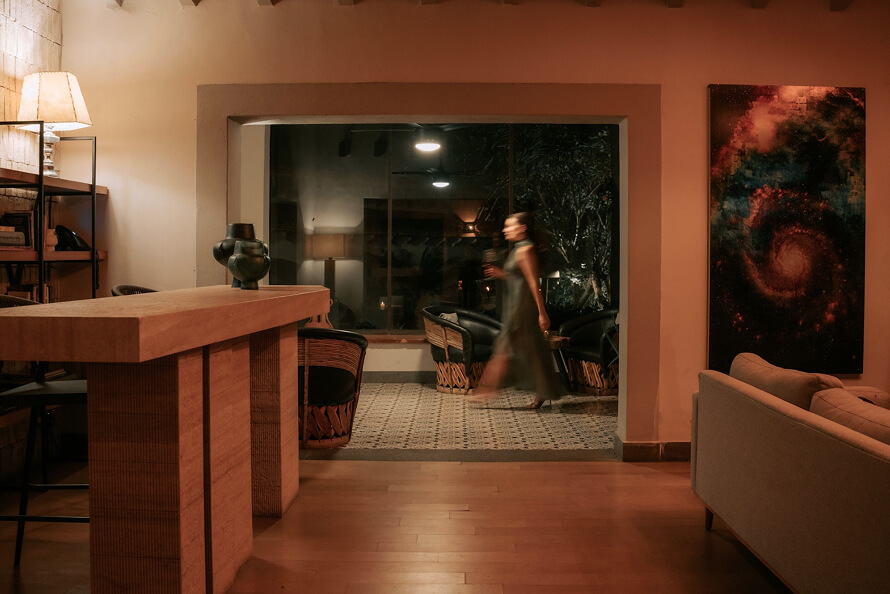 Interior cálido de una sala con luz tenue, una mujer caminando y un gran ventanal al fondo.