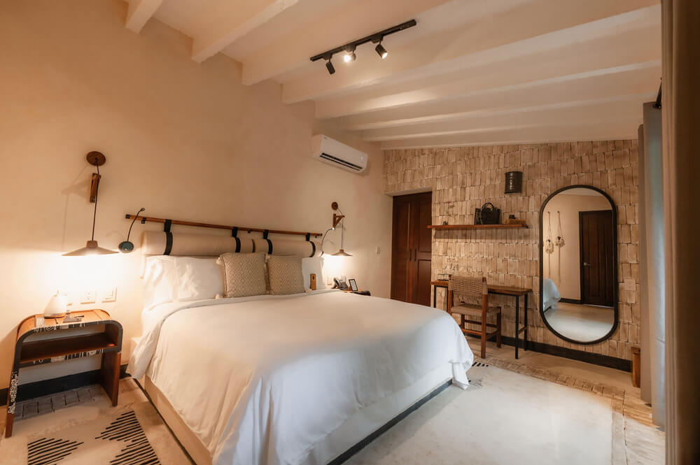 Dormitorio moderno con cama grande, ropa blanca, lámparas de pared, escritorio con silla y espejo ovalado.