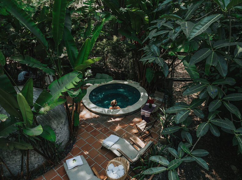 Persona disfrutando en un jacuzzi rodeado de plantas y vegetación en un espacio al aire libre con suelo de baldosas.