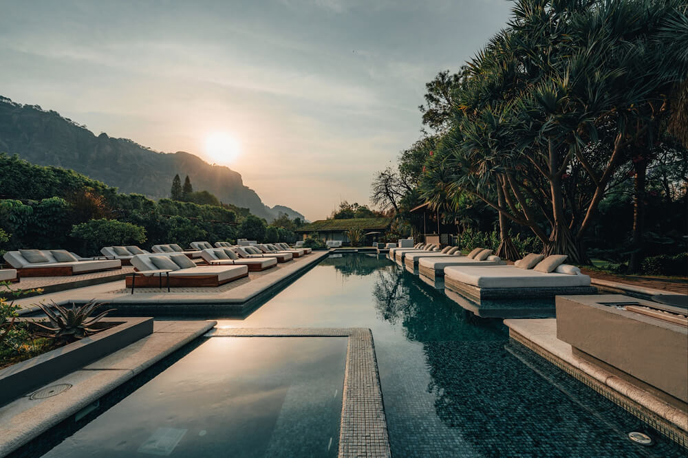 Piscina al aire libre con tumbonas acolchonadas a ambos lados y un atardecer detrás de montañas y árboles.