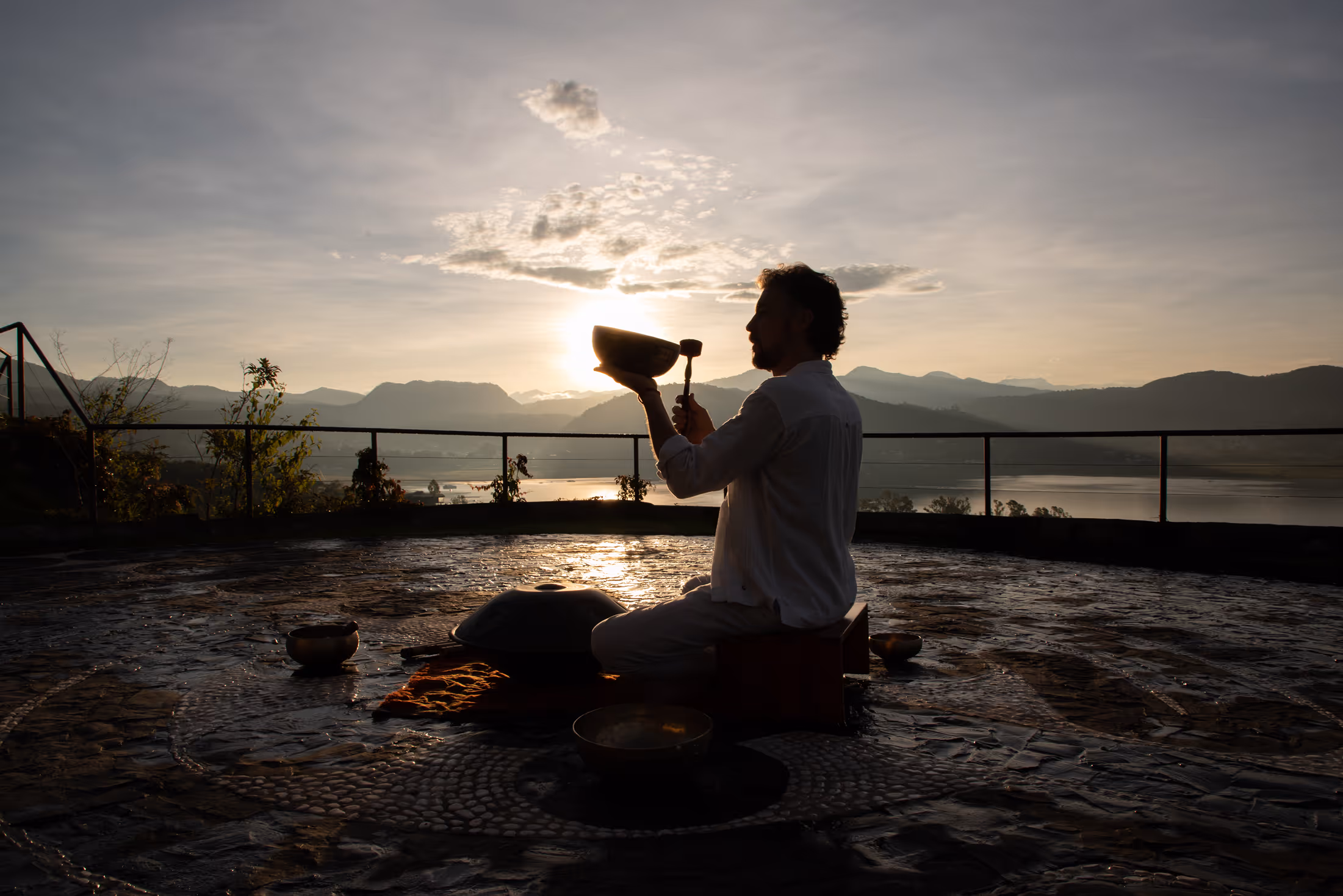 Persona sentada en posición de meditación al atardecer, sosteniendo un cuenco tibetano con un mazo, con montañas y un cuerpo de agua al fondo.