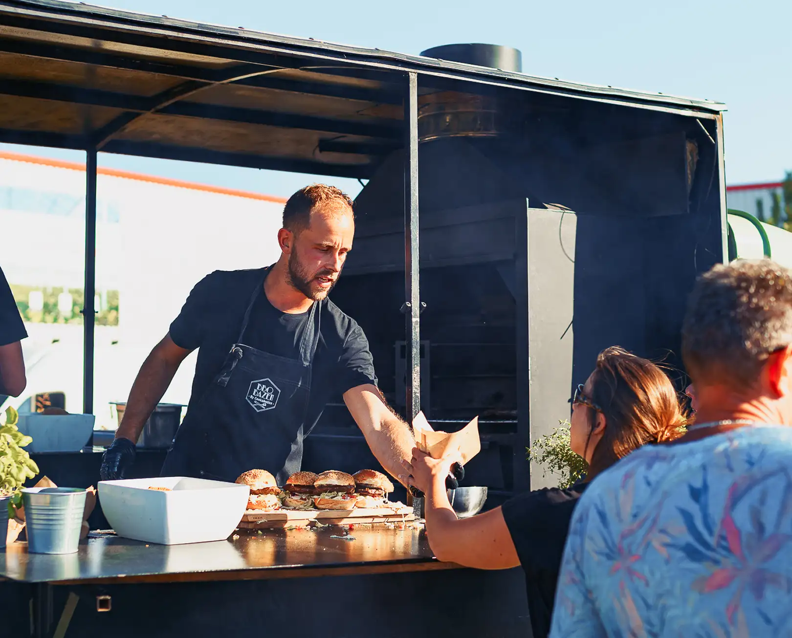 Man serveert hamburgers aan klanten bij een buitenbarbecuekraam op een zonnige dag.